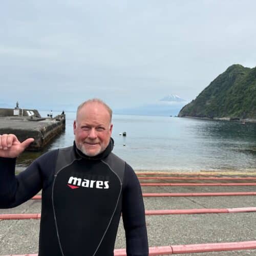 Man in wetsuit by ocean with mountain background.